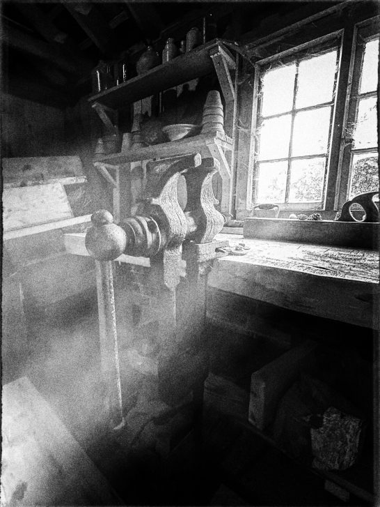 A Black and White image of a dusty workshop bench vice with sunlight beaming through a dirty glass window.