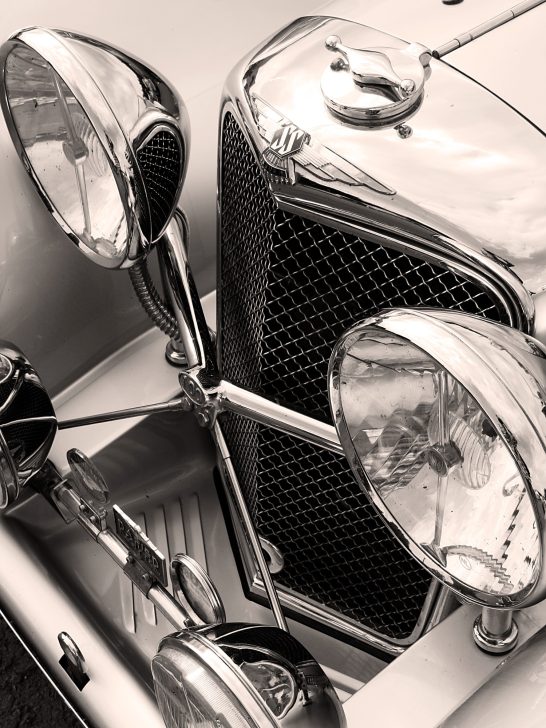 The image shows a close-up of the chrome radiator grille, winged "SS" badge, and large headlights of a vintage SS Jaguar 100 sports car.