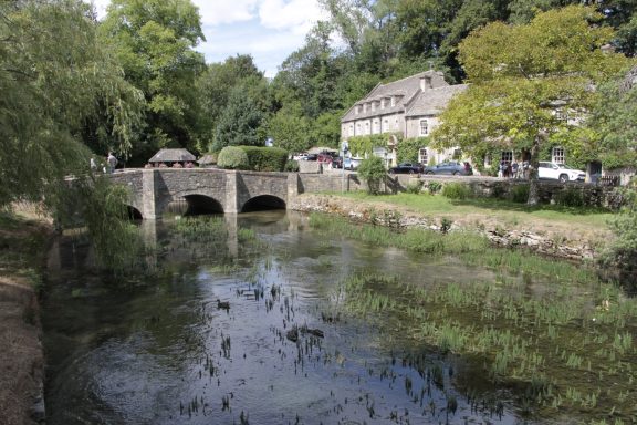This image shows the historic 3-arch stone bridge over the River Coln in Bibury, located in Gloucestershire in the Cotswolds.