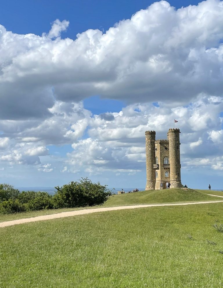 Photo of Broadway Tower in Worcestershire perched on a raised green plateau with a beautiful blue sky and fluffy white clouds